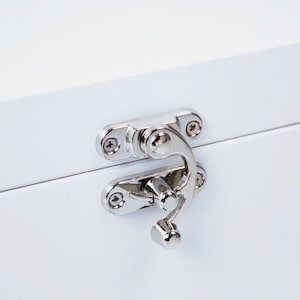 May include: Close-up of a white wooden box with a shiny silver latch. The latch is composed of two hinged plates and a curved handle, secured with small screws. The box's surface is smooth and reflective.