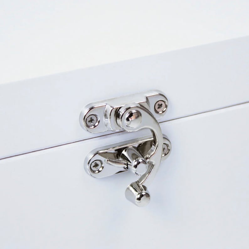 May include: Close-up of a white wooden box with a shiny silver latch. The latch is composed of two hinged plates and a curved handle, secured with small screws. The box's surface is smooth and reflective.