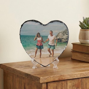 May include: Heart-shaped slate photo with a beach scene, held by a clear plastic stand. The photo shows a couple walking on the beach. The slate has a rough, dark edge, and is displayed on a wooden surface.