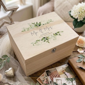 Könnte beinhalten: Eine hellfarbene Holzkiste mit Scharnierdeckel. Der Deckel ist mit den Worten "Mr & Mrs" und dem Datum "27th July 2023" sowie floralen Akzenten verziert. Die Kiste dient wahrscheinlich zur Aufbewahrung von Andenken.