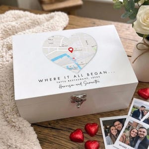 May include: A white keepsake box with a heart-shaped map design on the lid. The map has a red marker, and the text "WHERE IT ALL BEGAN..." is printed below. The box has a silver clasp and is surrounded by photos and heart-shaped chocolates.