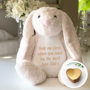 Op de afbeelding: Een witte pluche konijn met lange oren en een roze neus. Het konijn zit op een wit oppervlak en heeft de tekst "Hold me close when you need me the most Love Dad x" geborduurd op zijn borst. In de rechterbenedenhoek van de afbeelding bevindt zich een gouden hartvormige herinneringsdoos.