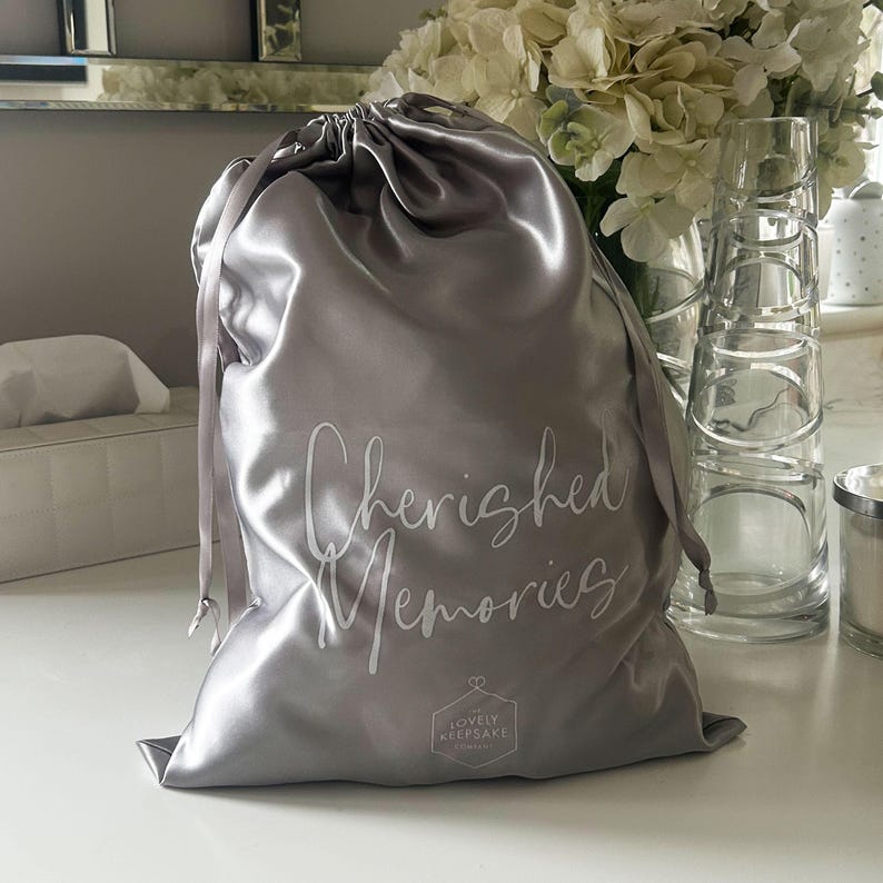 May include: A silver satin drawstring bag with the text "Cherished Memories" and a small logo that says "Lovely Keepsake" printed on the front.