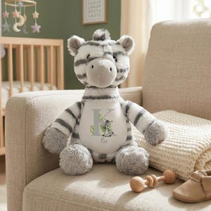 Op de afbeelding: Een pluche zebra met zachte wit-grijze gestreepte vacht. De zebra heeft een wit buikvlak bedrukt met een grote grijze letter K, een kleine illustratie van een babyzebra, groene bladeren en de naam Kai geschreven in een cursief lettertype.