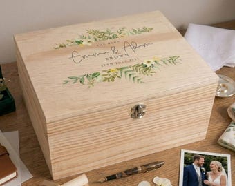 May include: A rectangular light-colored wooden keepsake box with a metal latch. The lid features a printed botanical design in green and yellow, with the text "The Barn", "Emma & Adam", "Brown", and "17th July 2023" in black script and print.
