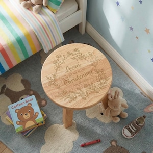 May include: A small, round wooden stool with a floral design and the words "Leoni Christening" engraved on the top. The stool has three legs and is surrounded by children's items, including a book and a stuffed toy.