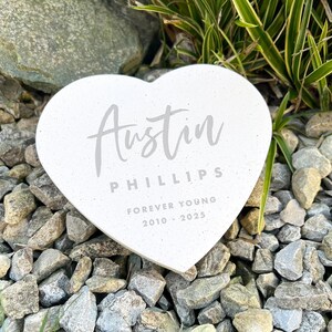 May include: A heart-shaped memorial stone with the name "Austin" and the last name "Phillips" in gray lettering. The text "Forever Young 2010-2025" is also inscribed. The stone is white and rests on a bed of small gray rocks.