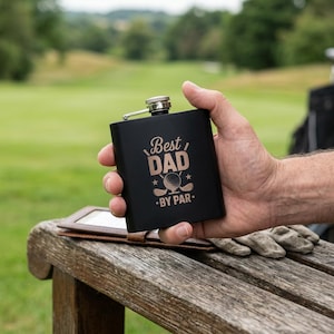 Könnte beinhalten: Ein mattschwarzer Metallflachmann mit einer Gravur, die den Text "Best DAD BY PAR" sowie einen Golfball und zwei Golfschläger zeigt. Der Flachmann hat einen silberfarbenen Schraubverschluss und wird vor einem unscharfen Golfplatz in der Hand gehalten.