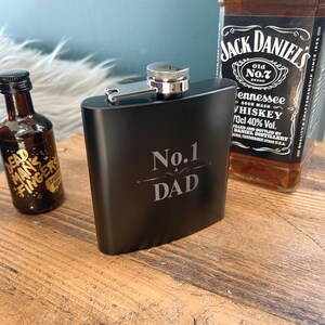 Può includere: Una fiaschetta nera con tappo e cerniera argentati, incisa con "No.1 DAD" in bianco. Una bottiglia di whisky Jack Daniel's e una piccola bottiglia di rum Dead Man's Fingers sullo sfondo, suggerendo un set regalo.