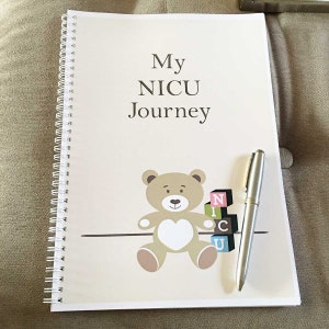May include: A beige journal with a spiral binding and the text "My NICU Journey" on the cover. A brown teddy bear with a white heart on its chest is holding a stack of colourful blocks that spell out "NICU".