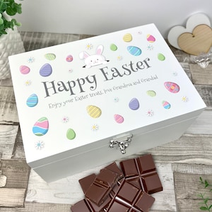 Puede incluir: Una caja de madera blanca para golosinas de Pascua con tapa con bisagras. La tapa está decorada con coloridos huevos de Pascua, margaritas y un conejito de dibujos animados. Las palabras "Felices Pascuas" están impresas en la tapa, junto con el mensaje "Disfruta de tus golosinas de Pascua, de la abuela y el abuelo."