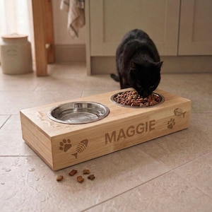 Puede incluir: Un soporte rectangular de madera clara para mascotas con dos cuencos circulares de acero inoxidable. El frente de la base de madera está grabado con el nombre MAGGIE, junto a pequeños iconos de huellas y espinas de pescado. Un gato negro come.