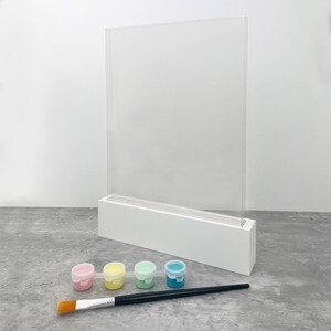 May include: A clear acrylic display stand with a white base, accompanied by four small paint pots in pink, yellow, green, and blue, and a paintbrush. The stand is designed to hold artwork or photos, perfect for creative projects.