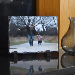 Puede incluir: Impresión fotográfica rectangular en pizarra con una imagen en color de una persona y un niño caminando por un sendero. La pizarra está soportada por dos soportes metálicos negros. El fondo incluye árboles y un jarrón.