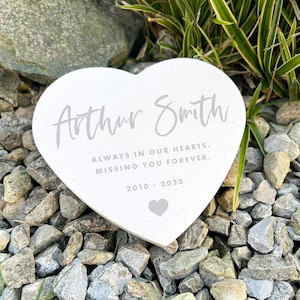 May include: A white heart-shaped memorial stone with the name "Arthur Smith" in script. The text "ALWAYS IN OUR HEARTS. MISSING YOU FOREVER." is below the name, along with the years 2010-2025 and a heart symbol. The stone rests on a bed of gray rocks.