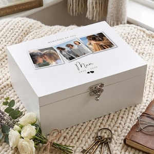 Könnte beinhalten: Eine weiße Holz-Andenkenbox mit Scharnierdeckel und Metallverschluss. Der Deckel zeigt drei gerahmte Fotos und den Text "MEMORIES OF YOU" und "Mum 1962-2026". Die Box liegt auf einer cremefarbenen Strickdecke.