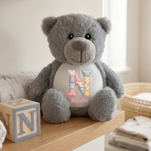 Può includere: Un orsacchiotto di peluche grigio con una toppa circolare bianca sul petto. La toppa presenta una grande lettera N in stile patchwork multicolore e il nome Natalie stampato in rosa sotto di essa. L'orso ha occhi scuri e un naso cucito.