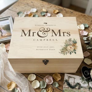 May include: A rectangular wooden keepsake box with a hinged lid and metal latch. The lid features the text "WEDDING MEMORIES", "Mr & Mrs", "CAMPBELL", "27TH JULY 2023", and "RIVERVALE BARN" in dark brown, alongside a floral illustration.