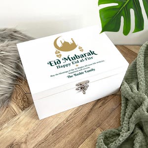 Op de afbeelding: Een witte houten kist met een zilveren sluiting. Het deksel is voorzien van de tekst "Eid Mubarak Happy Eid al-Fitr" in groen en goud, samen met een illustratie van een maan en moskee. De tekst "May the blessings of the Al-Mighty fill your life with joy, happiness and success. The Bashir Family" is ook aanwezig.
