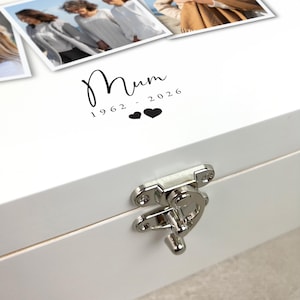 May include: A white wooden box with a silver clasp. The lid features the word "Mum" in a script font, along with the years "1962 - 2026" and two heart symbols. The box has a glossy finish and appears to be a keepsake box.
