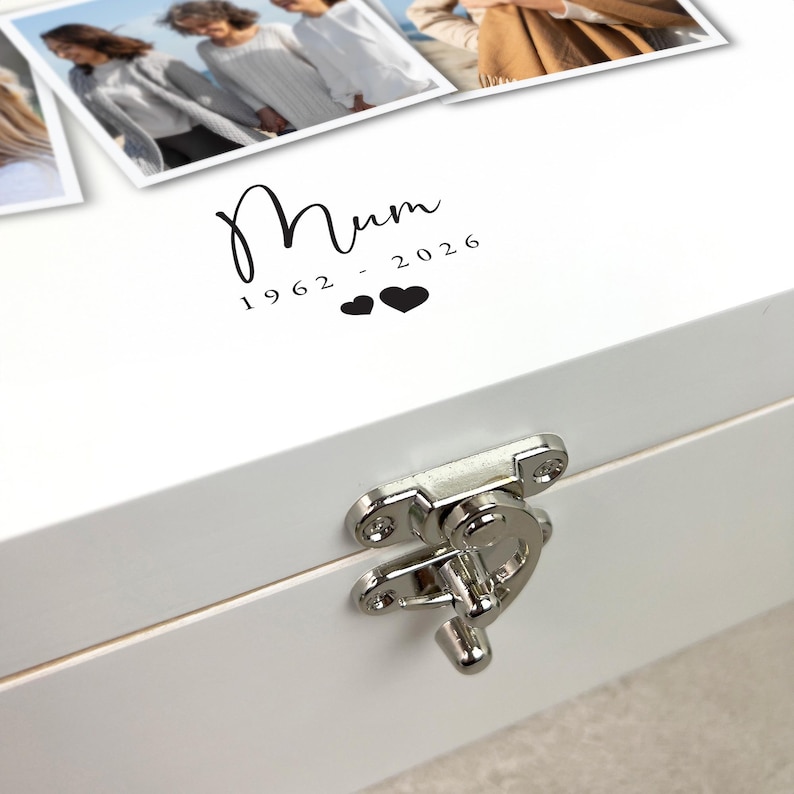 May include: A white wooden box with a silver clasp. The lid features the word "Mum" in a script font, along with the years "1962 - 2026" and two heart symbols. The box has a glossy finish and appears to be a keepsake box.