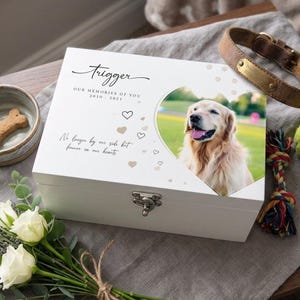 May include: A white wooden pet memorial box with the name "Trigger" in script. It features a heart-shaped photo frame with a golden retriever, text, and decorative hearts. A dog collar, toy, and flowers are nearby.