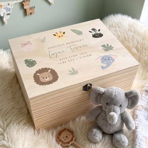 May include: A wooden keepsake box with animal illustrations and the text "Precious Memories Logan Turner 21st June 2024 7 LBS 9OZ 5.32AM". The box is decorated with a giraffe, lion, elephant, and other animal designs. A gray elephant plush toy and a wooden lion rattle are next to the box.