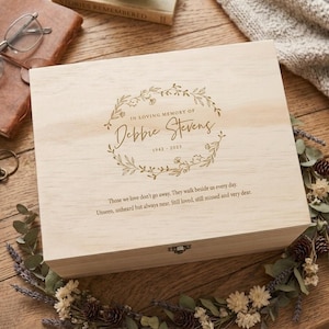 Può includere: Un cofanetto ricordo in legno chiaro con un design a ghirlanda floreale e la scritta "IN LOVING MEMORY OF Debbie Stevens 1942-2023." La scatola include anche un'iscrizione. Un oggetto decorativo per la commemorazione.