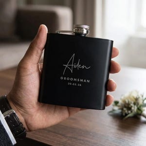 May include: A black hip flask with a silver cap, inscribed with the name "Aiden", the word "Groomsman", and the date "20.05.26" in white script. The flask is held in a hand, with a dark watch visible.