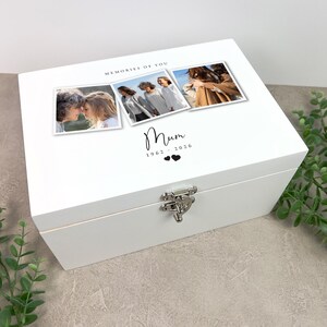 May include: A white wooden keepsake box with a silver clasp. The lid features three framed photos and the text "MEMORIES OF YOU", "Mum", and the years "1962-2026" with two heart symbols. The box is on a light gray surface.