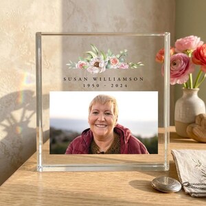 Könnte beinhalten: Ein Gedenkrahmen aus klarem Acrylblock mit einem gedruckten Aquarell-Blumenarrangement über dem Text SUSAN WILLIAMSON 1950 - 2024. Der Rahmen zeigt ein Foto einer lächelnden Person, die eine kastanienbraune Jacke und einen braunen Pullover trägt.