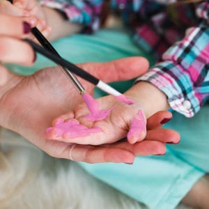 May include: A child's hand being painted with pink paint using a paintbrush. The hand is held by an adult, with a plaid shirt visible in the background. The image captures a moment of creative play.