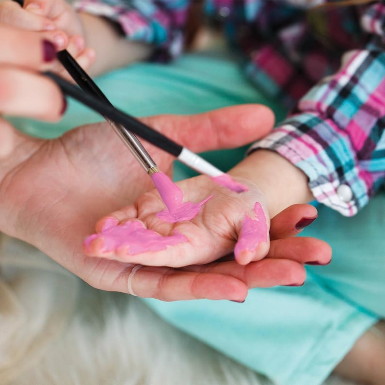 May include: A child's hand being painted with pink paint using a paintbrush. The hand is held by an adult, with a plaid shirt visible in the background. The image captures a moment of creative play.