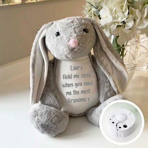 May include: Grey plush bunny with long ears, embroidered with the text "Laura Hold me close when you need me the most Grandma x". The bunny is sitting on a white surface. A white sound module is shown separately.