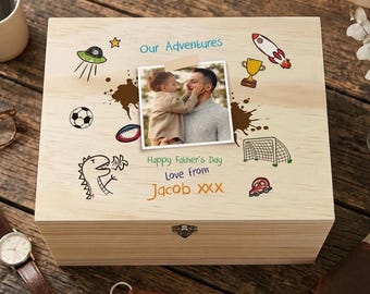 May include: A light-colored wooden keepsake box featuring a central photograph of a child and adult. The lid is decorated with colorful illustrations of a rocket, trophy, soccer ball, dinosaur, and UFO, with the text Our Adventures, Happy Father's Day, Love from Jacob XXX.