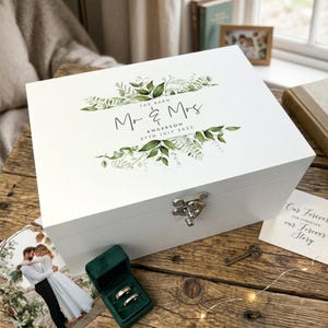 Peut inclure: Un coffret souvenir en bois blanc avec un fermoir argenté, orné d'illustrations botaniques et du texte "Mr & Mrs Anderson 27th July 2023". Une boîte à bagues en velours vert et une photo de mariage sont à proximité.