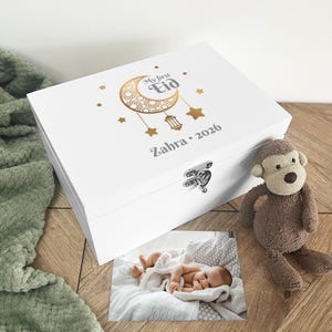 May include: A white wooden keepsake box with a silver clasp, featuring the text "My first Eid" and a crescent moon design. The box is personalized with a name and year. A plush monkey and a baby photo are next to the box.