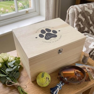 Peut inclure: Un coffret commémoratif en bois clair pour animaux de compagnie avec le texte "IN LOVING MEMORY OF" et "FOREVER IN OUR HEARTS". La boîte a une empreinte de patte et le nom "Marcus" avec les années 2011-2024. Une balle de tennis, un collier de chien et des photos sont à proximité.