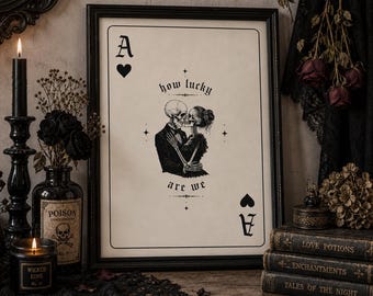 May include: A framed wall art print designed as an ace of hearts playing card featuring a black and white illustration of two skeletons embracing and the text how lucky are we centered on a cream background with black borders and corner symbols.