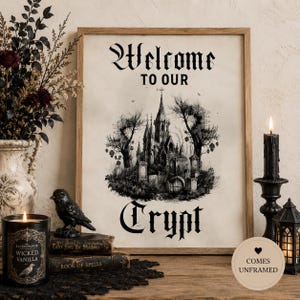 May include: A framed wall art print featuring a black and white illustration of a gothic castle surrounded by trees and tombstones. The text reads Welcome TO OUR Crypt in a stylised gothic font. The print is on a light cream background.