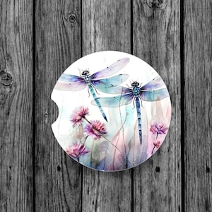 May include: Two blue and purple dragonflies with delicate wings are flying over a watercolor background of pink flowers and green foliage.