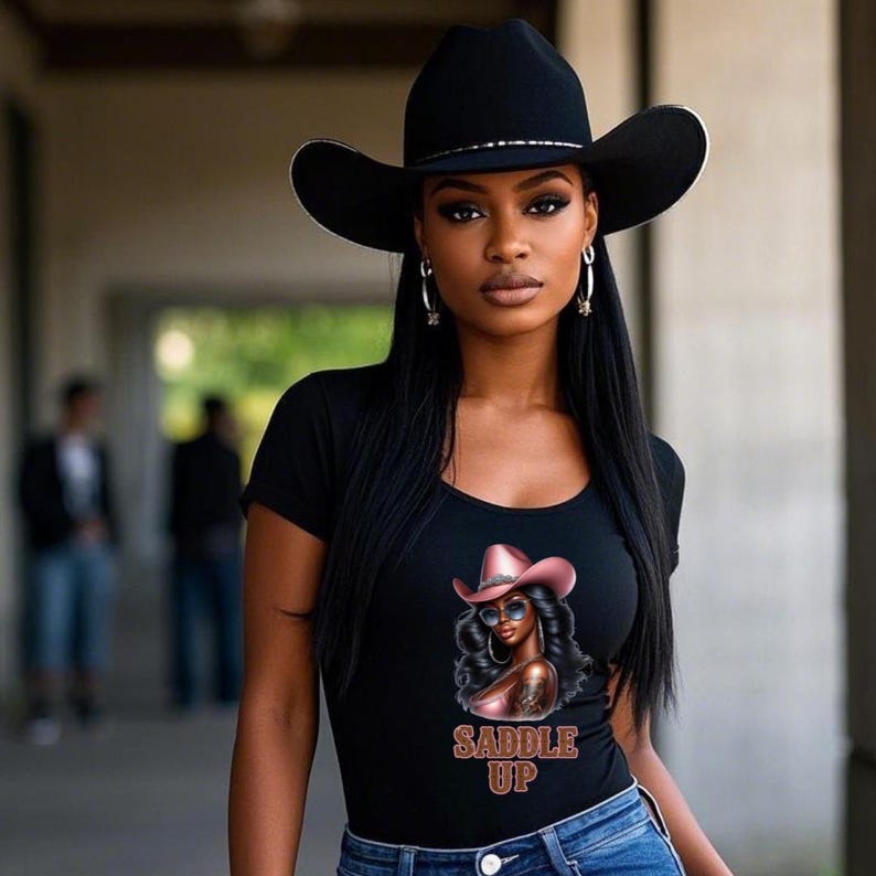 May include: A woman wearing a black cowboy hat and a black t-shirt with a graphic of a woman in a pink cowboy hat and the text "Saddle Up". She is also wearing blue jeans.