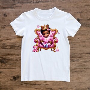 May include: White t-shirt with a graphic of a little girl wearing a crown and sitting on a pink throne. The girl is wearing a pink dress and has brown skin. There are butterflies and a teddy bear in the image.