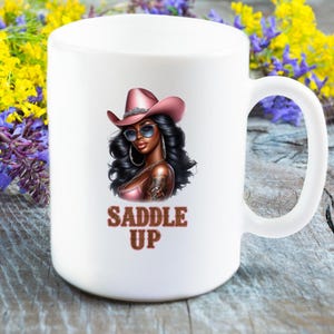 May include: White ceramic mug with a graphic of a woman wearing a pink cowboy hat and sunglasses. The text "Saddle Up" is printed in brown below the image.