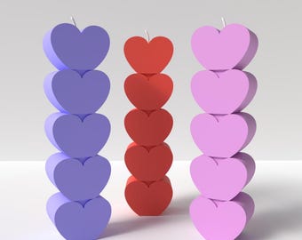 May include: Three stacked heart-shaped candles in different colors: purple, red, and pink. The candles are arranged in a row on a white surface.