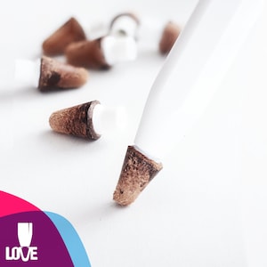 May include: A close-up of a white stylus with a brown wooden tip. The stylus is lying on a white surface with several small brown wooden objects scattered around it.