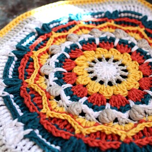 Op de afbeelding: Een kleurrijk gehaakt mandala met een wit centrum en concentrische ringen in rood, geel, turquoise en wit.