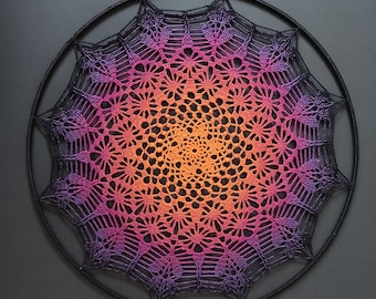 May include: A large, circular crochet mandala with a gradient of colors from purple to orange. The mandala is mounted on a black metal hoop.