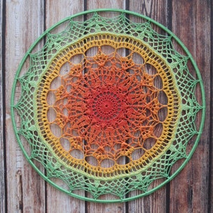 Puede incluir: Un mandala de ganchillo verde, amarillo y naranja con un marco de madera circular. El mandala tiene un patrón repetitivo de puntadas intrincadas.