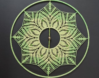 Puede incluir: Un atrapasueños de crochet verde y blanco con una pieza central de cuentas. El atrapasueños es circular y tiene un diseño delicado e intrincado.